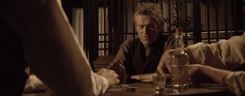 Movie still from “Dead for a Dollar” (2022), directed by Walter Hill – A man sitting at a wooden table with a glass of wine; Medium shot, Over the shoulder angle