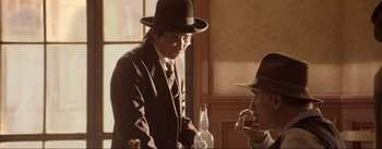 Movie still from “Dead for a Dollar” (2022), directed by Walter Hill – A man in a suit and a hat standing in front of a bottle; Medium shot, Over the shoulder angle