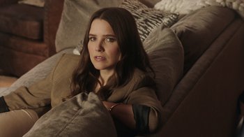 Movie still from “Deborah” (2022), directed by Noga Pnueli – A woman sitting on top of a couch in a living room; Medium shot, Over the shoulder angle