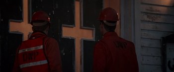 Movie still from “Die Hard 2” (1990), directed by Renny Harlin – Two men in red shirts and hard hats standing in front of a cross; Medium shot, Over the shoulder angle
