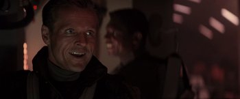 Movie still from “Die Hard 2” (1990), directed by Renny Harlin – A man smiling at the camera with another man in the background; Close Up shot, Over the shoulder angle