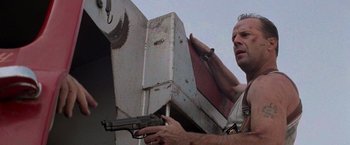 Movie still from “Die Hard with a Vengeance” (1995), directed by John McTiernan – A man holding a gun in his right hand while standing in front of an object; Close Up shot, Low angle