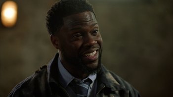 Movie still from “Die Hart” (2023), directed by Eric Appel – A person wearing a suit and tie smiling; Close Up shot, Over the shoulder angle