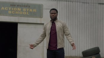 Movie still from “Die Hart” (2023), directed by Eric Appel – A man standing in front of a building with his arms outstreched; Medium shot, Over the shoulder angle