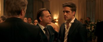 Movie still from “Die in a Gunfight” (2021), directed by Collin Schiffli – Two men in suits talking to each other in front of a crowd; Close Up shot, Over the shoulder angle