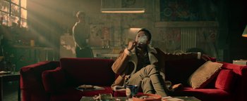 Movie still from “Die in a Gunfight” (2021), directed by Collin Schiffli – A man sitting on top of a red couch smoking a cigarette; Medium shot, Low angle