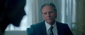 Movie still from “Die in a Gunfight” (2021), directed by Collin Schiffli – A man in a suit and tie sitting in front of a window; Close Up shot, Over the shoulder angle