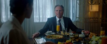 Movie still from “Die in a Gunfight” (2021), directed by Collin Schiffli – A man sitting at a dinner table with food on the table; Medium shot, Over the shoulder angle