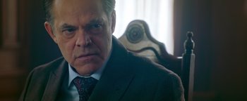 Movie still from “Die in a Gunfight” (2021), directed by Collin Schiffli – A man in a suit and tie sitting in a chair; Close Up shot, Over the shoulder angle