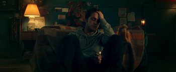 Movie still from “Die in a Gunfight” (2021), directed by Collin Schiffli – A man sitting on a couch holding a lit cigarette; Medium shot, High angle