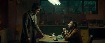 Movie still from “Die in a Gunfight” (2021), directed by Collin Schiffli – Two men sitting at a table in front of a lamp; Medium shot, Low angle