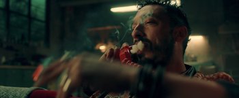Movie still from “Die in a Gunfight” (2021), directed by Collin Schiffli – A man smoking a cigarette while holding a pipe in his mouth; Close Up shot, Low angle