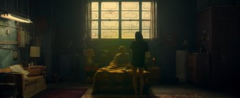 Movie still from “Die in a Gunfight” (2021), directed by Collin Schiffli – A woman standing in front of a bed with a man sitting on it; Wide shot, High angle