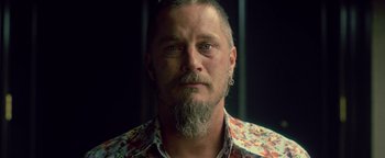 Movie still from “Die in a Gunfight” (2021), directed by Collin Schiffli – A person with a beard wearing a floral shirt; Close Up shot, Over the shoulder angle