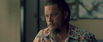 Movie still from “Die in a Gunfight” (2021), directed by Collin Schiffli – A man with a long beard and a flower shirt; Close Up shot, Over the shoulder angle