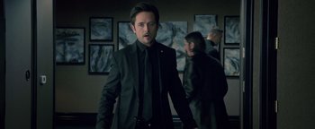 Movie still from “Die in a Gunfight” (2021), directed by Collin Schiffli – A man in a black suit and black tie standing in front of a group of people; Medium shot, Over the shoulder angle