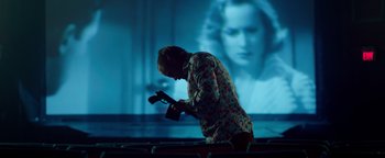 Movie still from “Die in a Gunfight” (2021), directed by Collin Schiffli – A man holding a camera in front of a projection of a woman; Medium shot, Low angle