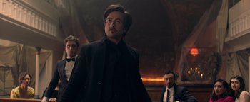 Movie still from “Die in a Gunfight” (2021), directed by Collin Schiffli – A man in a suit and tie standing in front of two other men; Medium shot, Low angle