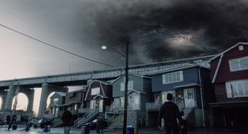 Movie still from “War of the Worlds” (2005), directed by Steven Spielberg – People walking on the sidewalk in front of a row of houses under a cloudy sky; Extreme Wide shot, Low angle