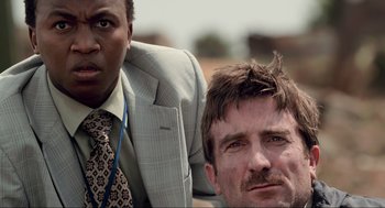 Movie still from “District 9” (2009), directed by Neill Blomkamp – A man and a man in a suit sitting next to each other; Close Up shot, Over the shoulder angle