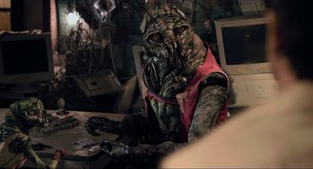 Movie still from “District 9” (2009), directed by Neill Blomkamp – A person in a costume sitting at a desk; Medium shot, Over the shoulder angle