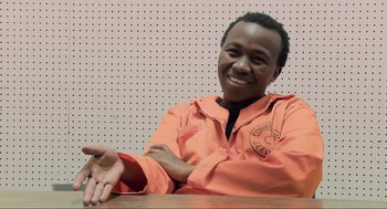 Movie still from “District 9” (2009), directed by Neill Blomkamp – A man sitting at a table in an orange prison uniform; Close Up shot, Over the shoulder angle
