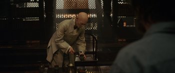 Movie still from “Doctor Strange” (2016), directed by Scott Derrickson – A bald man in a white suit is holding a cup; Medium shot, Over the shoulder angle