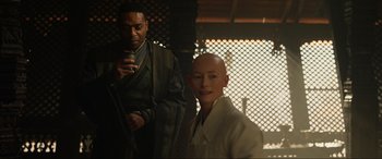 Movie still from “Doctor Strange” (2016), directed by Scott Derrickson – A man standing next to a woman with a cell phone in front of him; Medium shot, Over the shoulder angle