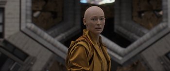Movie still from “Doctor Strange” (2016), directed by Scott Derrickson – A woman with a bald head is standing in front of a building; Close Up shot, Over the shoulder angle
