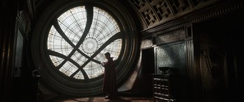 Movie still from “Doctor Strange” (2016), directed by Scott Derrickson – A man in a red robe standing in front of a stained - glass window; Wide shot, Over the shoulder angle