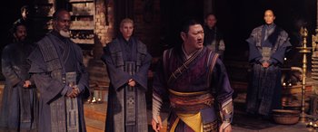 Movie still from “Doctor Strange in the Multiverse of Madness” (2022), directed by Sam Raimi – Two men dressed in traditional chinese garb stand next to each other in front of a brick wall; Medium shot, Over the shoulder angle