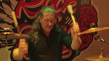 Movie still from “Doll House” (2022), directed by Marla Ancheta – A man with long hair is holding a drum stick; Medium shot, Low angle