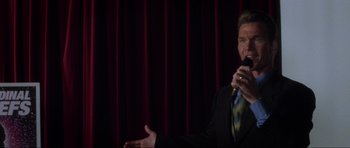 Movie still from “Donnie Darko” (2001), directed by Richard Kelly – A man in a black suit is speaking into a microphone; Medium shot, Over the shoulder angle