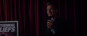 Movie still from “Donnie Darko” (2001), directed by Richard Kelly – A man holding a microphone while standing in front of a red curtain; Close Up shot, Over the shoulder angle