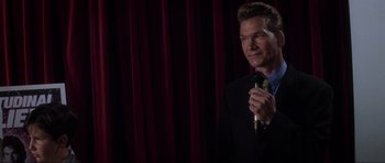 Movie still from “Donnie Darko” (2001), directed by Richard Kelly – A man holding a microphone in front of a red curtain; Close Up shot, Over the shoulder angle