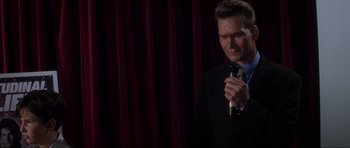 Movie still from “Donnie Darko” (2001), directed by Richard Kelly – A man in a suit and tie holding a microphone in his hand; Close Up shot, Low angle