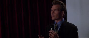 Movie still from “Donnie Darko” (2001), directed by Richard Kelly – A man in a suit and tie holding a microphone; Close Up shot, Over the shoulder angle