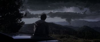 Movie still from “Donnie Darko” (2001), directed by Richard Kelly – A man sitting on the hood of a car in front of a cloudy sky; Extreme Wide shot, Over the shoulder angle