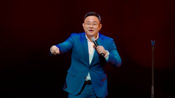 Movie still from “Dr Jason Leong: Ride with Caution” (2023), directed by Jason Leong – A man in a blue suit is holding a microphone; Medium shot, Low angle