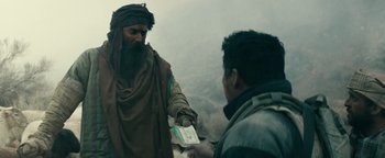 Movie still from “12 Strong” (2018), directed by Nicolai Fuglsig – A man in a brown robe is handing a card to another man in a green jacket; Medium shot, Over the shoulder angle