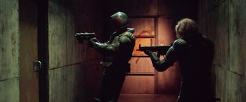 Movie still from “Dredd” (2012), directed by Pete Travis – Two people in a room with guns on a wall; Wide shot, Low angle