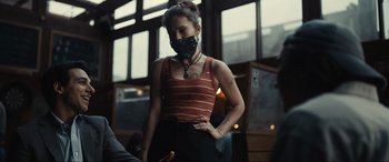 Movie still from “Dumb Money” (2023), directed by Craig Gillespie – A woman wearing a black face mask standing next to a window; Medium shot, Low angle