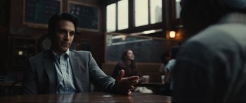 Movie still from “Dumb Money” (2023), directed by Craig Gillespie – A man sitting at a table in front of other people; Medium shot, Over the shoulder angle