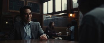 Movie still from “Dumb Money” (2023), directed by Craig Gillespie – A man sitting at a table in front of other people; Medium shot, Over the shoulder angle