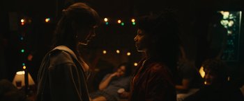 Movie still from “Dumb Money” (2023), directed by Craig Gillespie – Two women are standing in a room with lights in the background; Medium shot, Over the shoulder angle