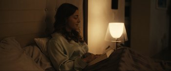 Movie still from “Dumb Money” (2023), directed by Craig Gillespie – A woman sitting in a chair reading a book; Medium shot, High angle