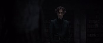 Movie still from “Dune” (2021), directed by Denis Villeneuve – A man in a black suit standing in a dark room; Medium shot, Over the shoulder angle