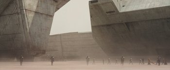 Movie still from “Dune” (2021), directed by Denis Villeneuve – A group of people walking in the sand near a large concrete structure; Extreme Wide shot, Low angle