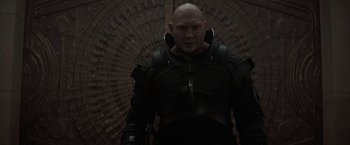 Movie still from “Dune” (2021), directed by Denis Villeneuve – A bald man in a black outfit standing in front of a brick wall; Medium shot, Over the shoulder angle