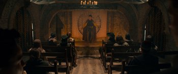 Movie still from “Dungeons & Dragons: Honor Among Thieves” (2023), directed by John Francis Daley – People are sitting in chairs in front of a stage with a man on it; Wide shot, High angle
